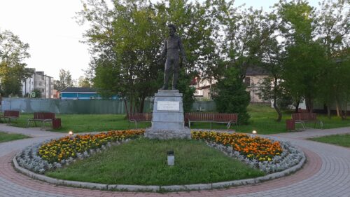 A photograph features a metal statue of a man on an elevated platform in a grassy circle in the center of a park. Orange and yellow flowers surround the platform.
