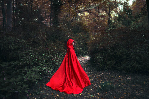 A photograph features a person in a red hooded cape standing in a dark forest, looking back at the viewer.