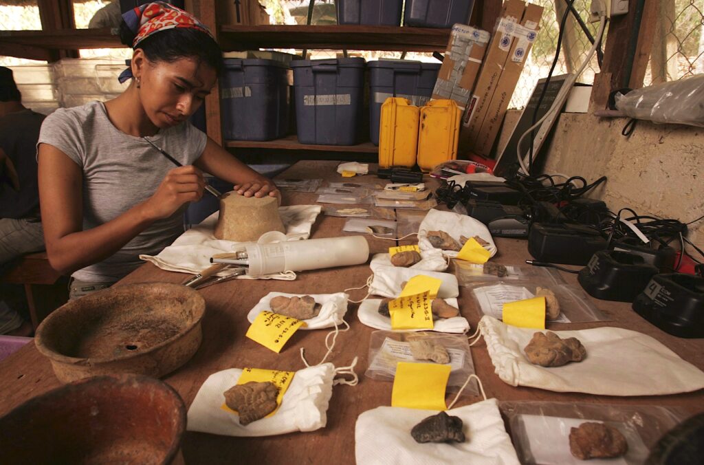 A person with black hair wearing a gray T-shirt sits at a table brushing a cylindrical clay object that stands next to several stone fragments labeled with yellow tags.