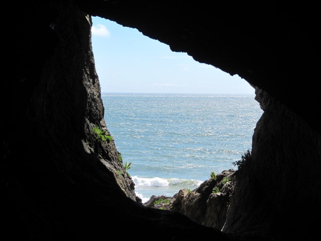 From inside a dark cave, a photograph features a rocky tear-shaped cave opening looking out on blue seawater.