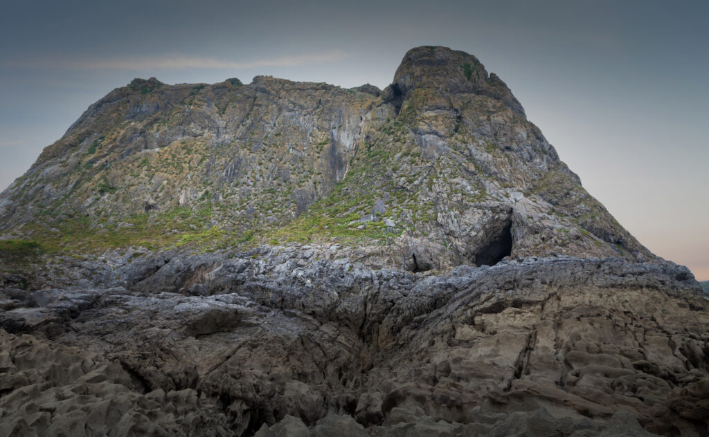 A photograph features a rocky landscape that ascends into a large mountain. A small teardrop-shaped opening is visible on the mountain’s right side.