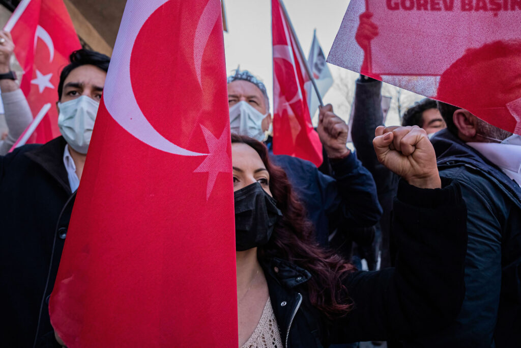 A photograph features a crowd of people wearing face masks and holding up red flags with a white star and crescent moon on them. Several people are also raising fists.