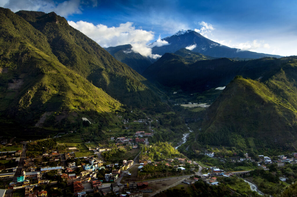 A photograph features a widescale view of a village’s buildings and roads with huge green mountains flanking its far side and a blue sky with white clouds above.