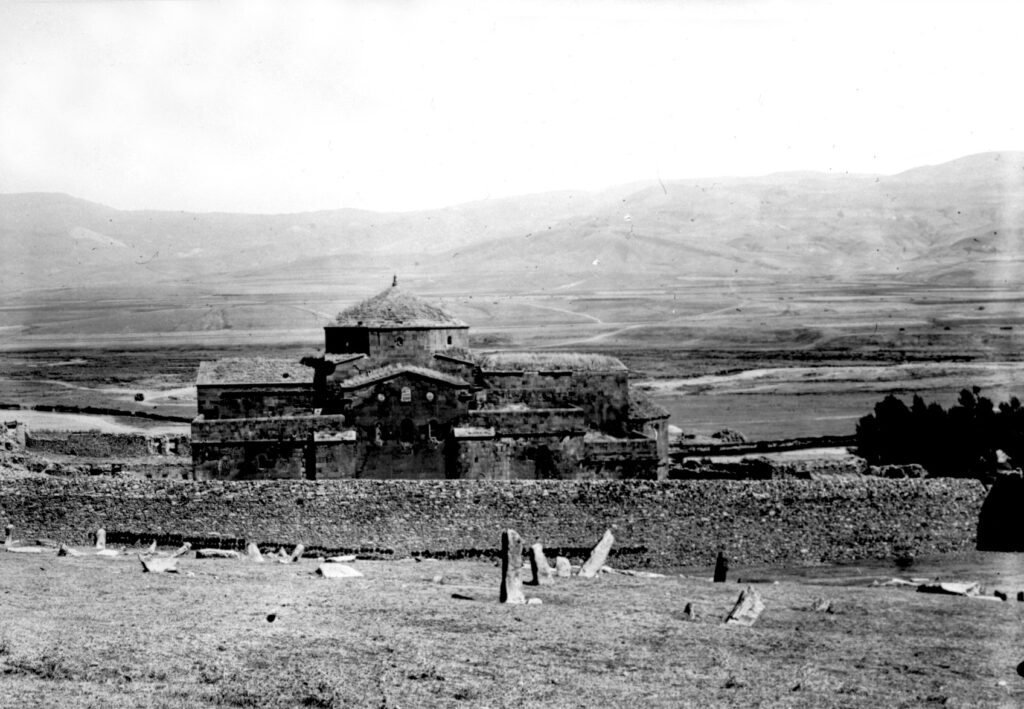 A black-and-white photograph features a large brick building surrounded by a stone wall in an expansive field. Rolling hills and a distant mountain range fill the background.