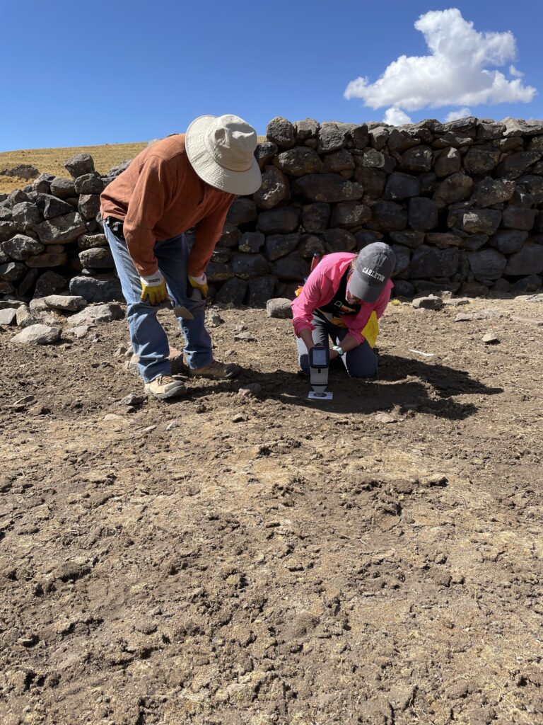 A photograph shows two people wearing brimmed hats, blue jeans, and boots. One kneels, holding a white instrument to the dirt ground, as the other stands and looks on.