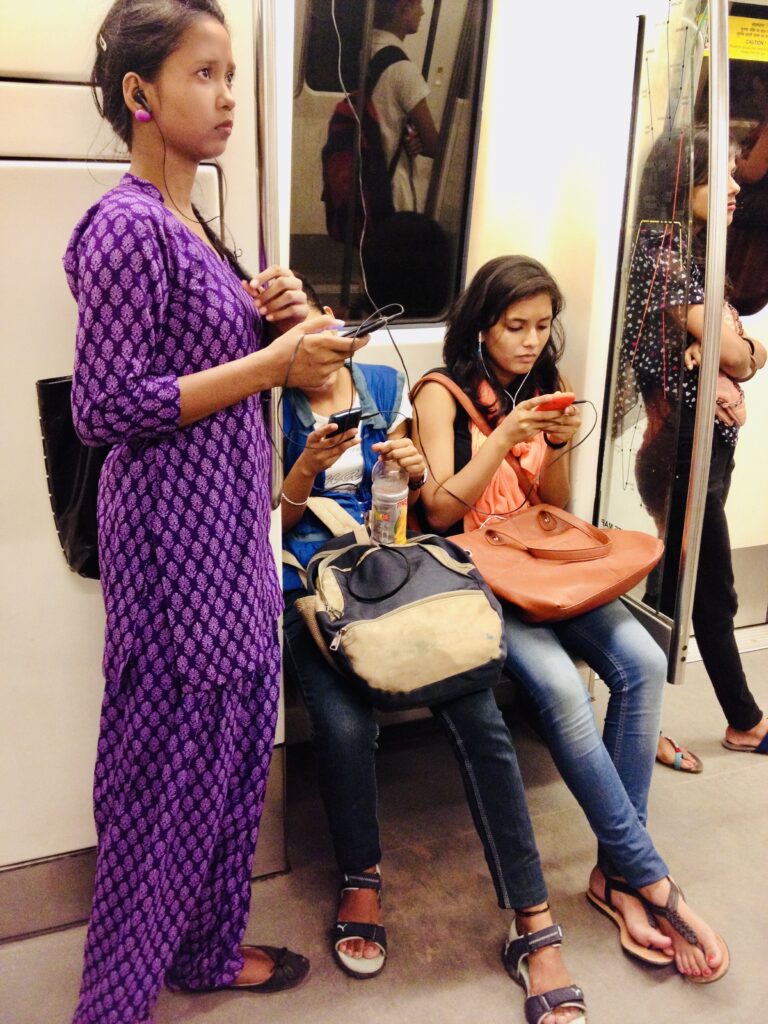 Two women sit in a metro car and use their cellphones. Another woman stands beside them with headphones in her ears.