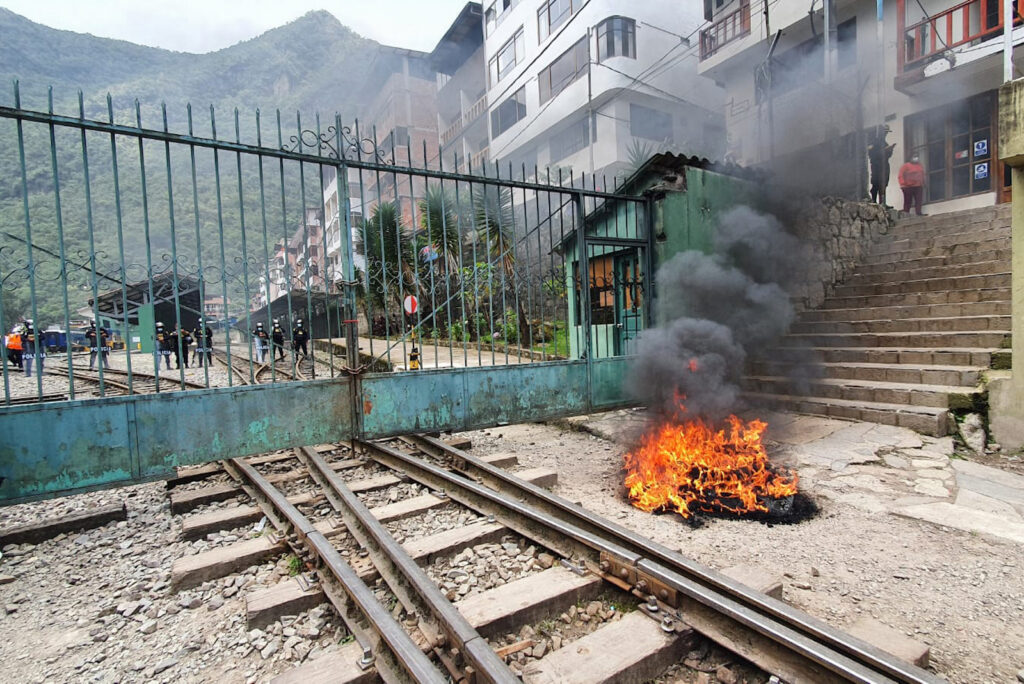 A photograph captures a small fire burning in front of a closed gate and next to a building, with a handful of masked people in the distance. Tall mountains are visible on the horizon.