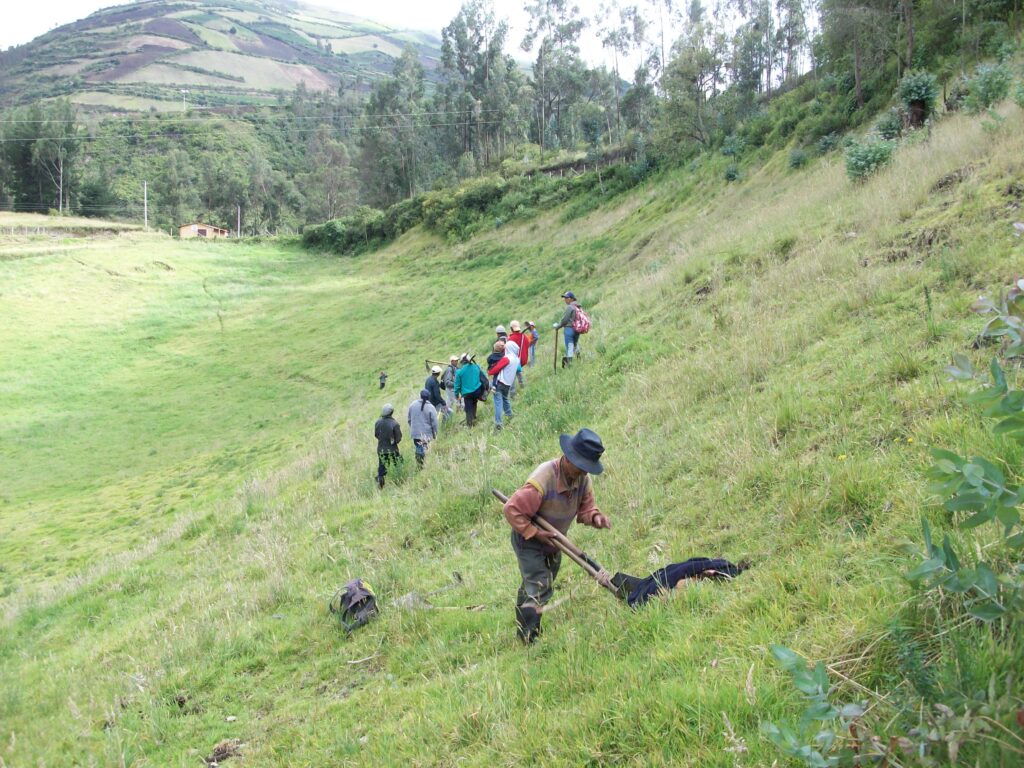 A photograph features several people wearing hats, jackets, and long pants gathered on a sloping grassy hillside. A person in the foreground holds a wooden stick to the earth.