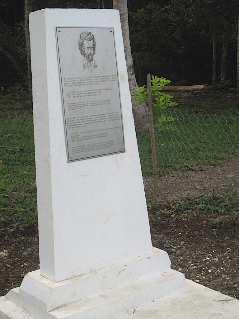 A photograph features a tall white tombstone on a dirt plot with a silver plaque on it that has a person’s face etched on top and text underneath.
