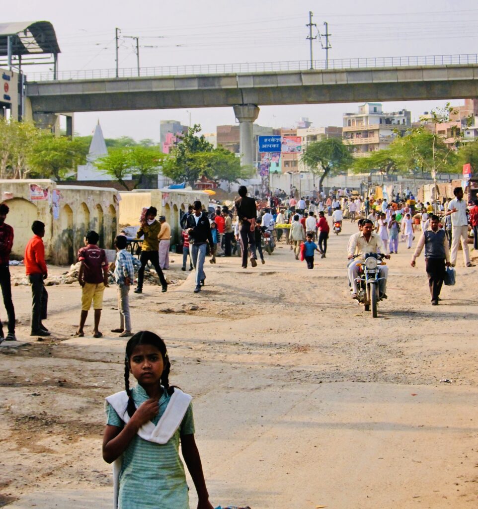 A photograph features a dirt street with many people walking or riding bikes below an elevated train track.