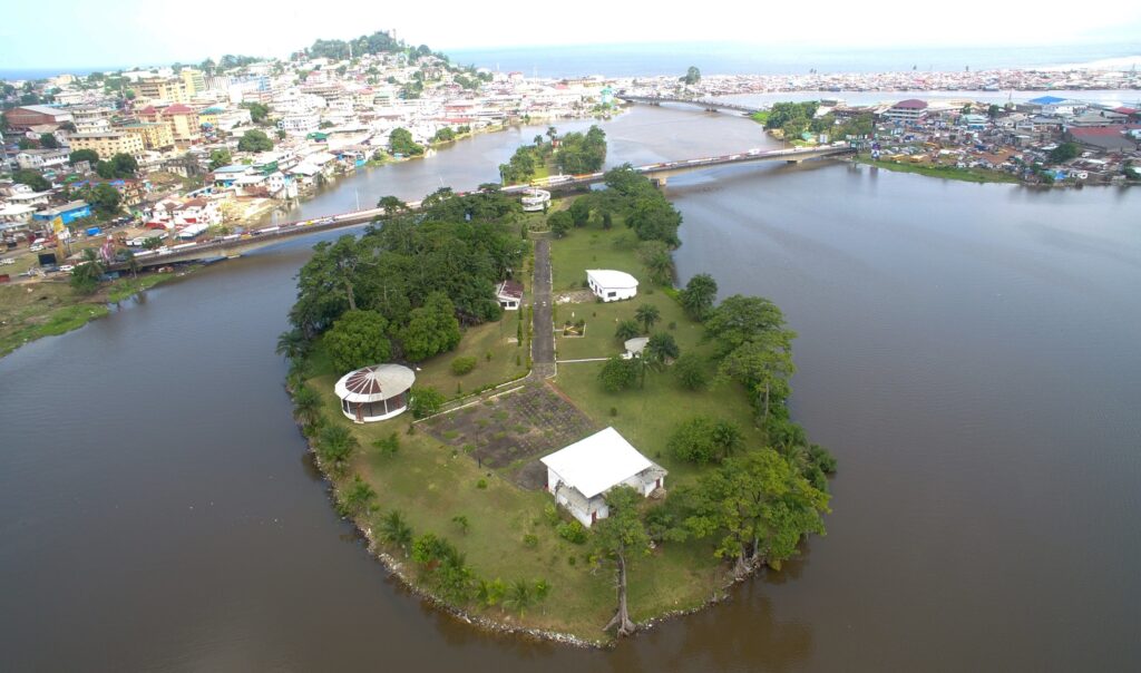 An aerial photograph features a grassy green island with trees and a few white buildings on it. Connected to the island by a long bridge, a shore full of buildings spans the background.