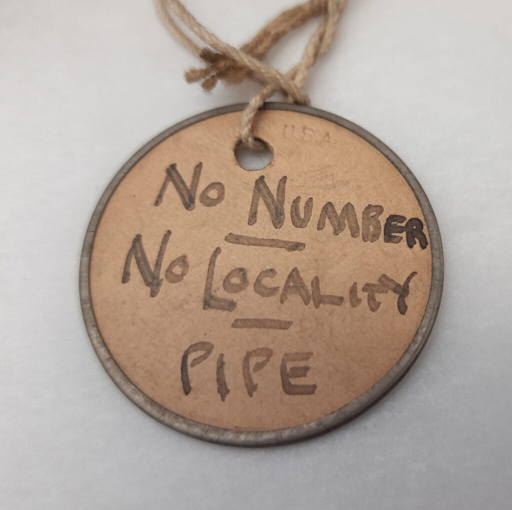 A cardboard tag tied with rope is labeled with the words “No Number, No Locality, Pipe” written in black marker.