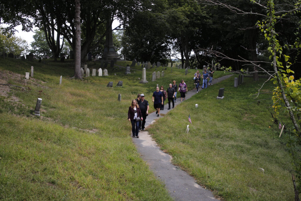 A group of people walk down a narrow concrete path flanked by lawns and gravestones.