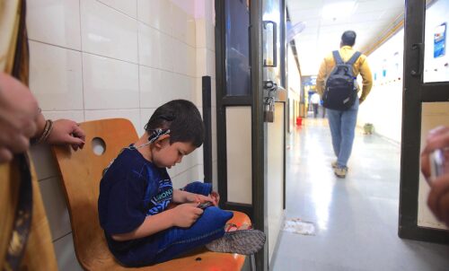 A photograph features a child with a cochlear implant sitting on a wooden chair at the end of a hallway. They are looking down at a handheld device they are using.