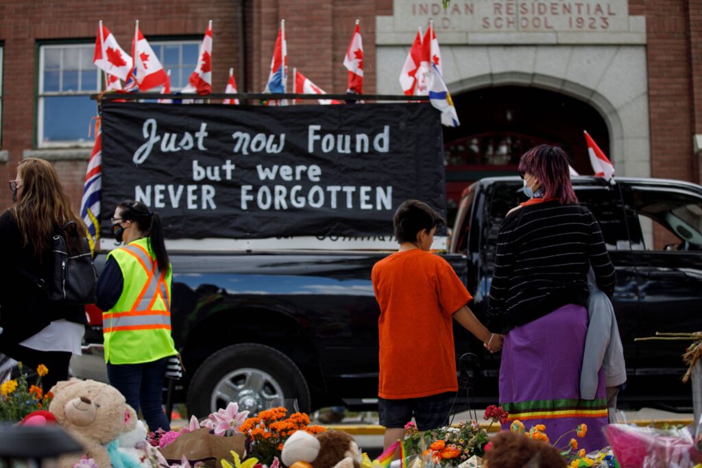 A photograph shows an adult and child watching a black truck pass in front of a memorial of flowers and stuffed animals. The truck has several Canadian flags flying from it and carries a black banner with white text that reads, “Just now found but were never forgotten.”