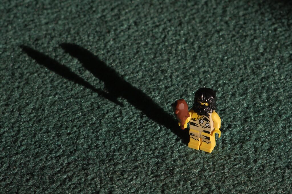 A close-up photograph features a yellow Lego toy shaped like a caveman holding a wooden club posed in front of a shadow it casts.
