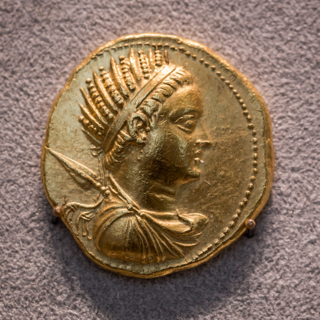 A gold coin shows a person’s profile from the shoulders up, with a spearhead etched in behind.