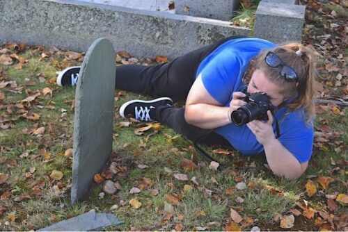 Lying on their side, a person with a blonde ponytail wears a blue shirt and black jeans and points a camera at a gravestone.