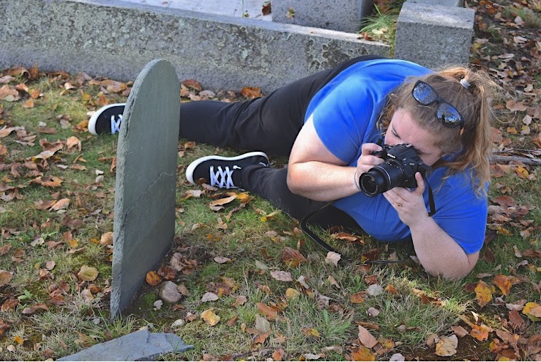 Lying on their side, a person with a blonde ponytail wears a blue shirt and black jeans and points a camera at a gravestone.