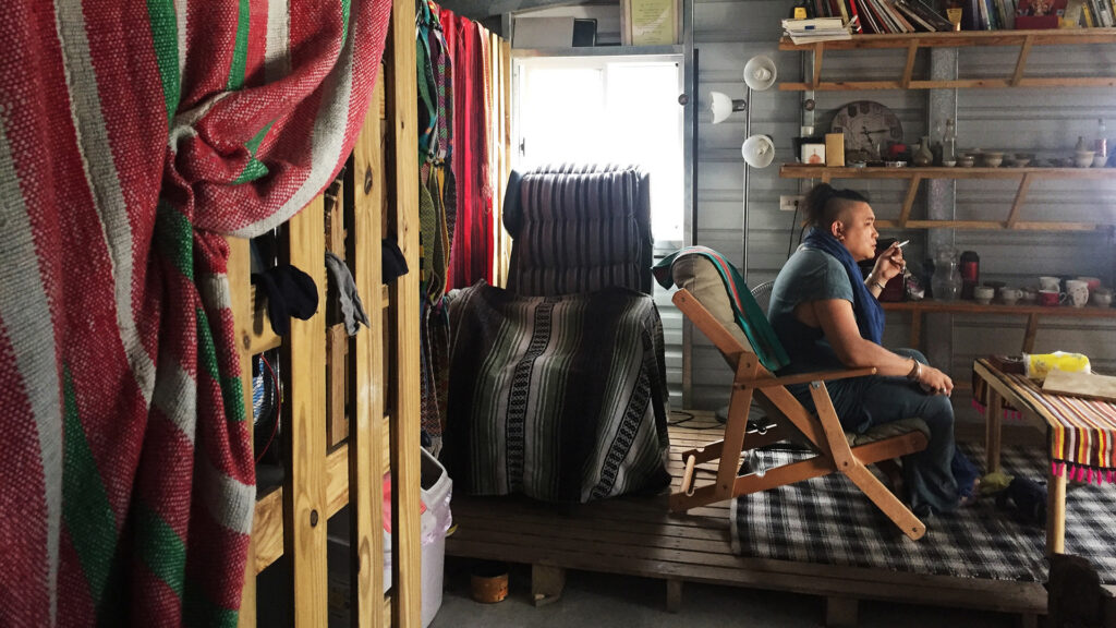 A person smokes a cigarette while sitting in a chair in a room with a plaid carpet, a large table, and wooden shelves.