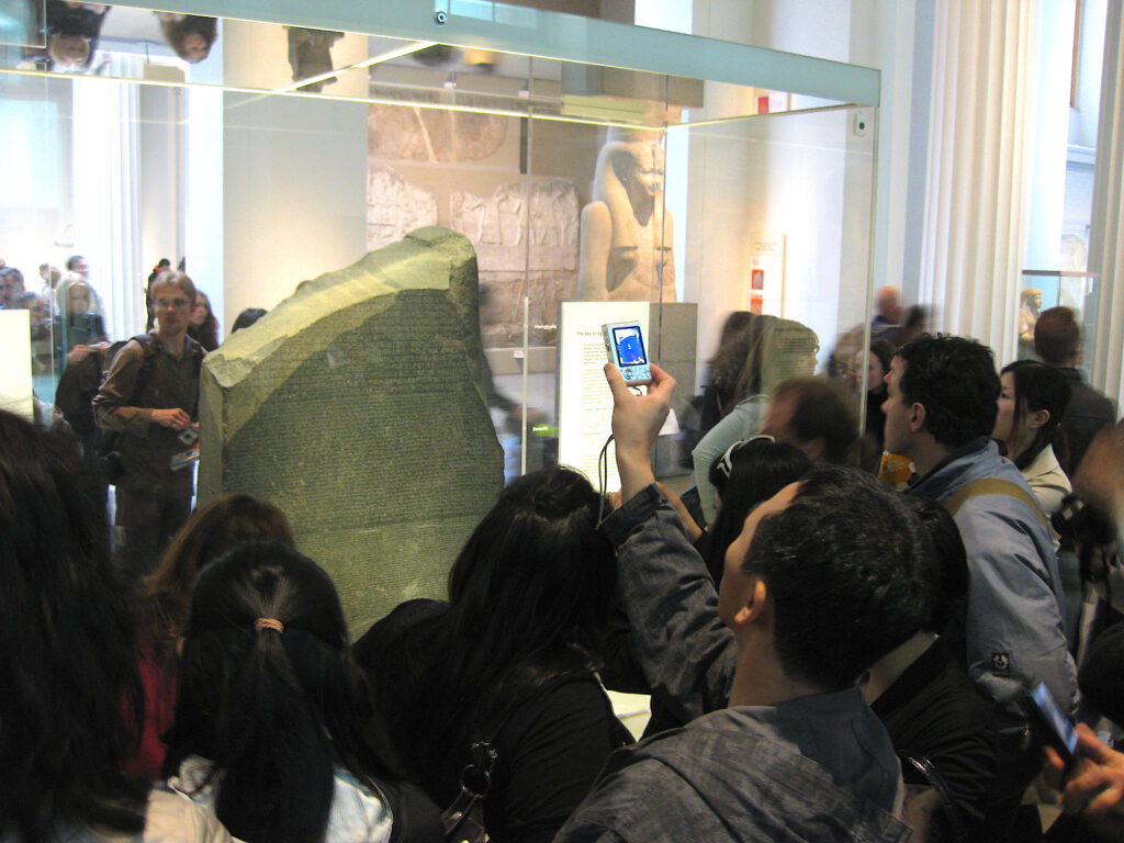 A crowd of people—some taking pictures with their cellphones—stand in front of a clear display case that houses a large gray stone.