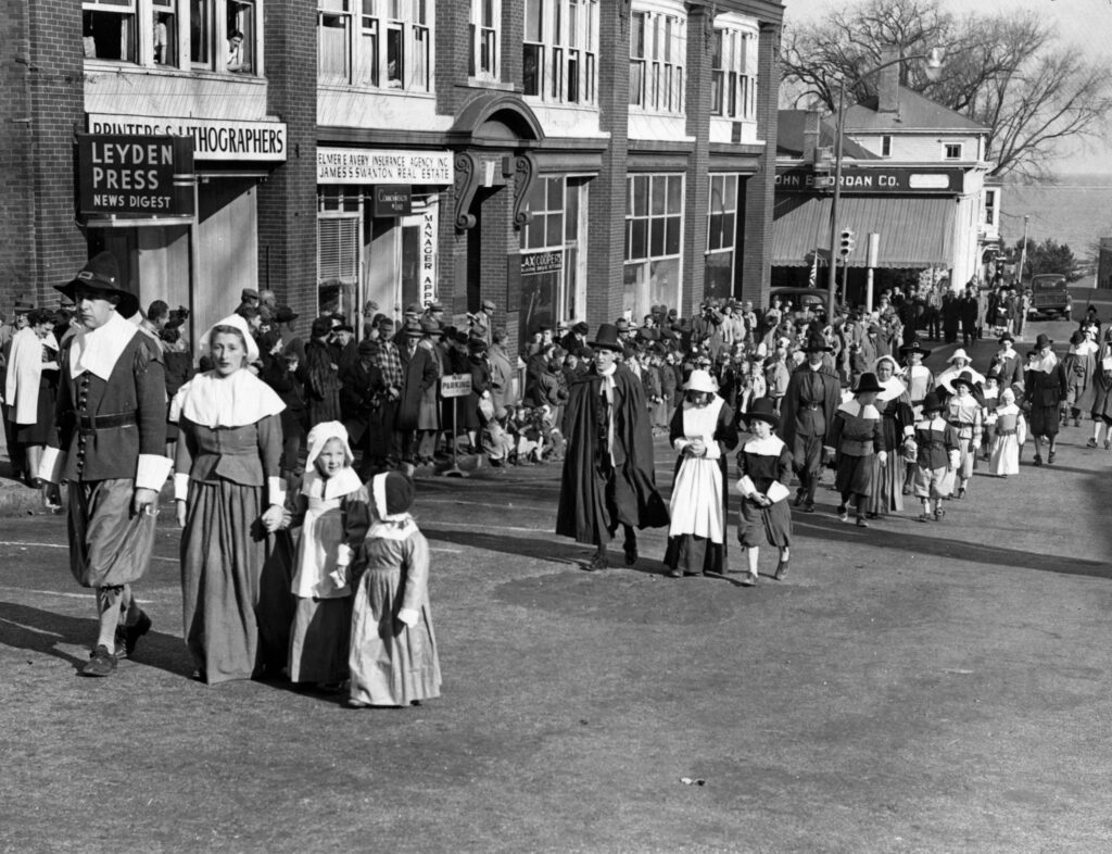 A black-and-white photograph features a town square full of people wearing collared shirts or dresses and white head coverings.