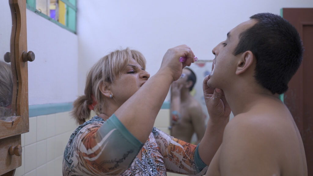A photograph features a person with straight blonde hair helping another person with short black hair to brush their teeth in a bathroom.