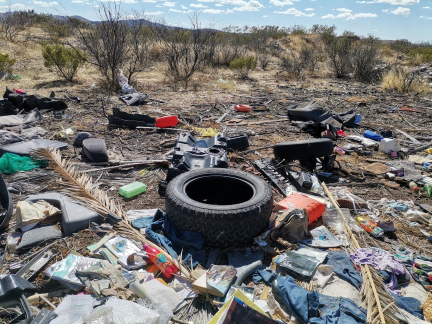 A photograph centers on a large dirt plot filled with garbage such as rubber tires, magazines, bottles, and a few large machine parts.