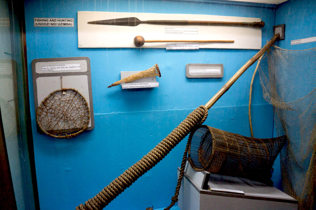 A photograph features several tools—nets, a spear, a stick with a spherical ball on the end, and a reed cage—in a glass case mounted on a blue wall.