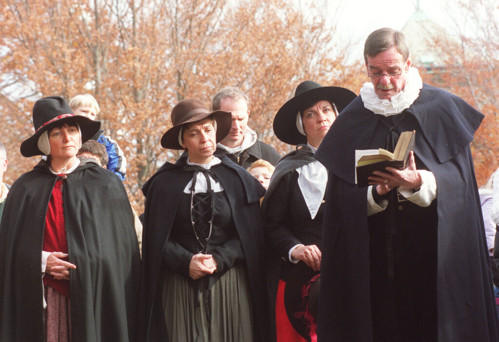 Three people dressed in black cloaks and brimmed hats stand with their hands crossed behind a hatless gray-haired person wearing a similar cloak.