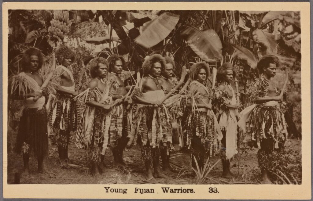 A sepia-tone photograph features a group of shirtless people in a forest wearing ornate loincloths and headdresses. Several carry wooden sticks.