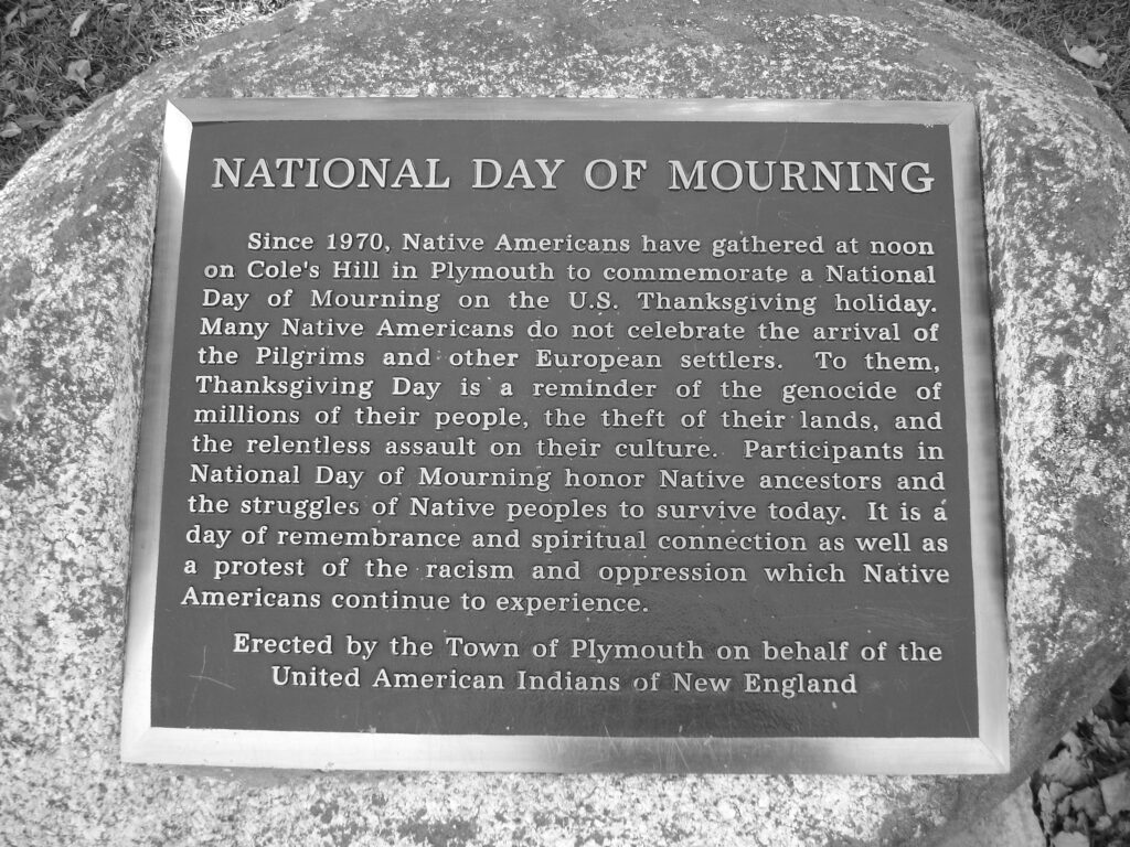 A close-up photograph features a large stone embedded with a plaque that reads, “National Day of Mourning. Since 1970, Native Americans have gathered at noon on Cole’s Hill in Plymouth to commemorate a National Day of Mourning on the U.S. Thanksgiving holiday. Many Native Americans do not celebrate the arrival of Pilgrims and other European settlers. To them, Thanksgiving Day is a reminder of the genocide of millions of their people, the theft of their lands, and the relentless assault on their culture. Participants in National Day of Mourning honor Native ancestors and the struggles of Native peoples to survive today. It is a day of remembrance and spiritual connection as well as a protest of the racism and oppression which Native Americas continue to experience. Erected by the Town of Plymouth on behalf of the United American Indians of New England.”