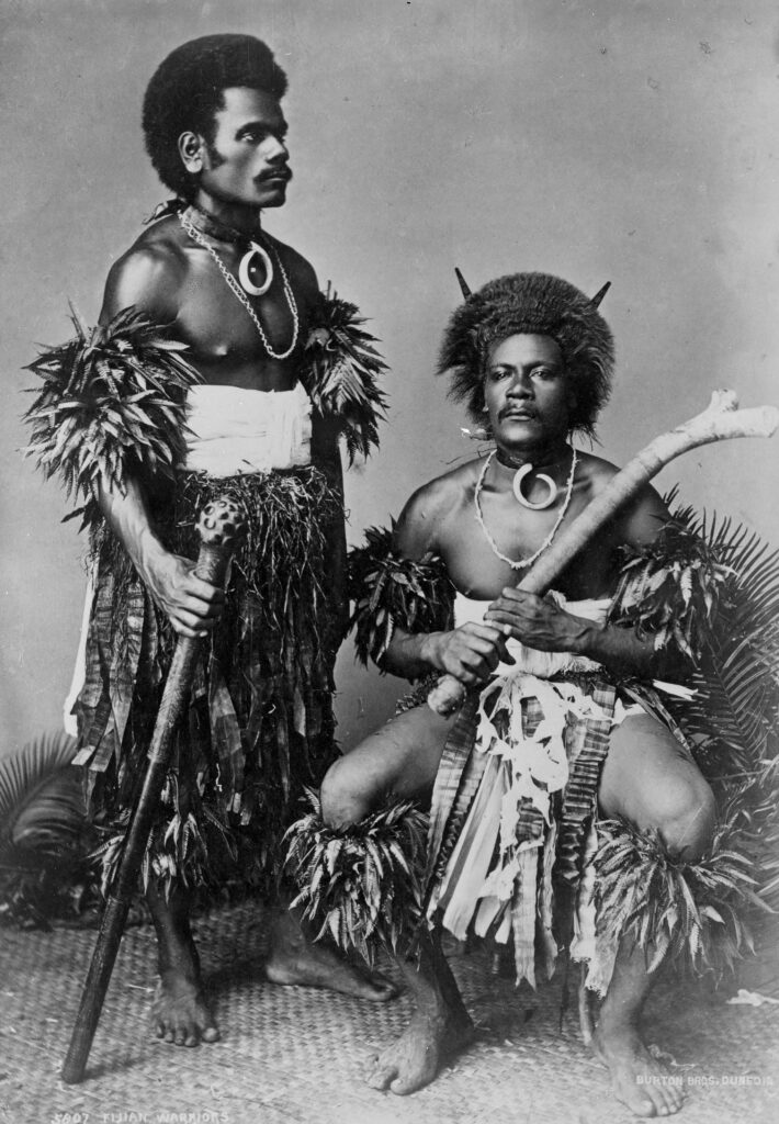 A black-and-white portrait features two people—one seated, and one standing—wearing ornate loincloths and headdresses. Both hold wooden sticks.
