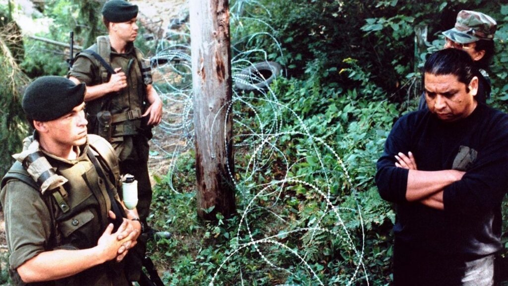 A photograph features three people in green military uniforms and one person dressed in all black standing in a forest on opposite sides of a wire fence.