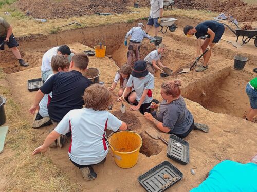 Several adults and children dig in a cleared area of brown soil with rectangular holes. Some dig with shovels and trowels, while others look on. Several buckets lie about.