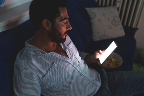 A man with black hair and a beard sits on a sofa in the dark and looks at the illuminated screen of a cellphone he holds in his left hand.