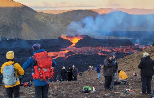 Several people wearing puffy hooded coats, beanies, windbreakers, and backpacks watch lava and smoke pour out of a volcano on the horizon.