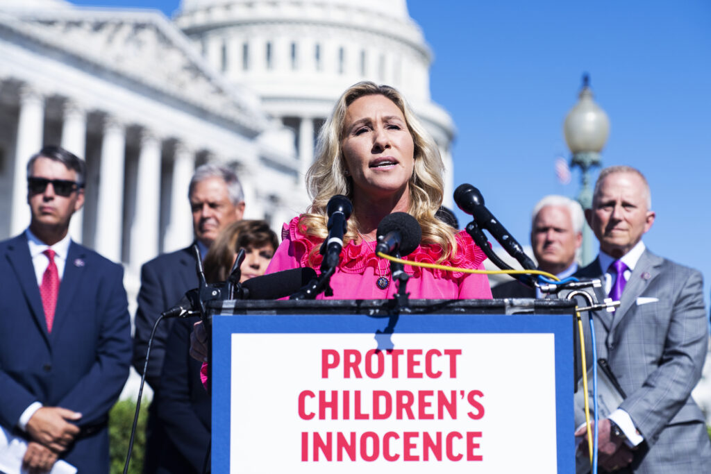 A person with blonde hair wearing a pink blazer stands at a podium with several microphones on it and a sign that reads, “Protect Children’s Innocence.” Several people in suits stand behind, with a large white domed building in the background.