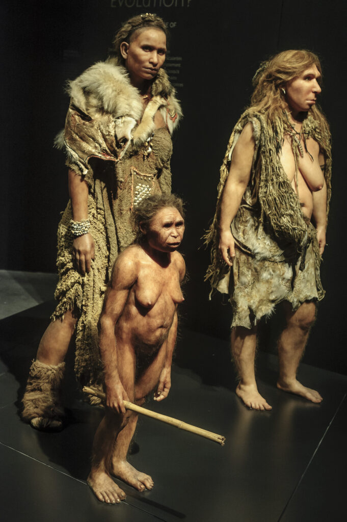 Three human-like statues stand in triangular orientation. The two in the back are tall and wear furs while the one in front wears no clothes and holds a short stick.