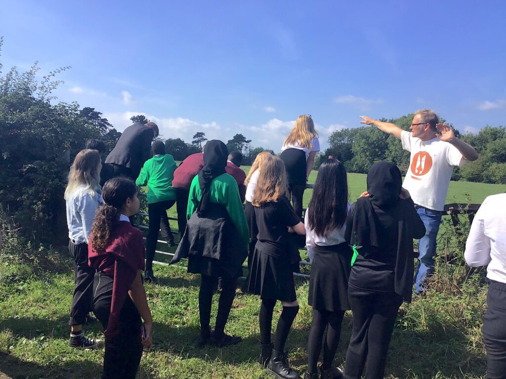 Children gather around a person wearing a white T-shirt and jeans and pointing to the right.