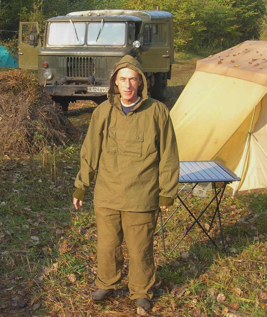 A person wearing black boots and a green hooded windbreaker and pants stands on a grassy area with a yellow tent and green jeep in the background.