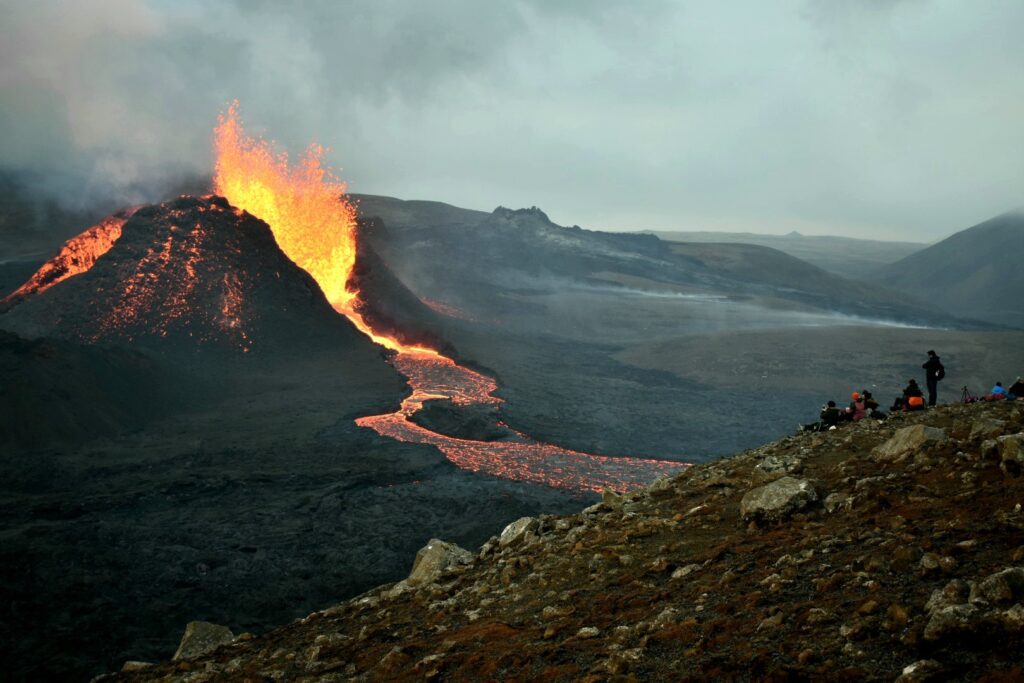 Lava erupts from a volcano on the far horizon as a group of people watch from rocky terrain in the foreground.