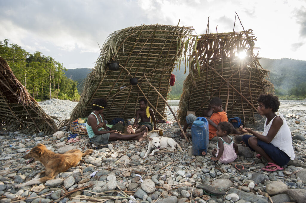 A group of adults, children, and two dogs sit on a bed of rocks under slanted walls made of wooden sticks and green plants.