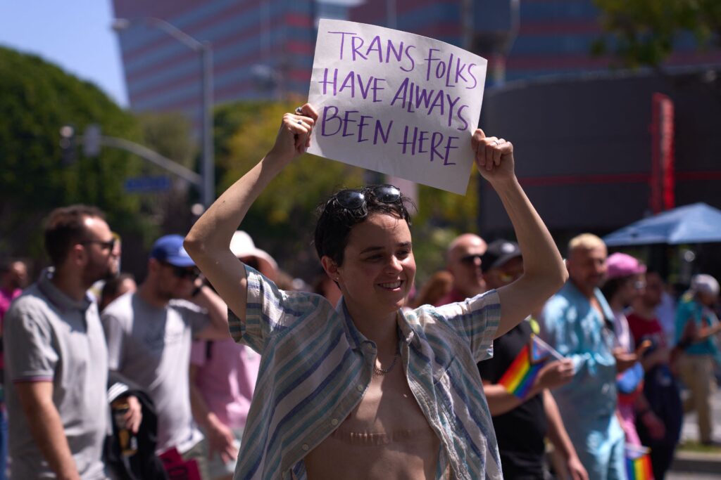 In front of a crowd of people carrying rainbow flags, a person with short brown hair is wearing an open-collared shirt and holding a sign that reads, “Trans Folks Have Always Been Here.”