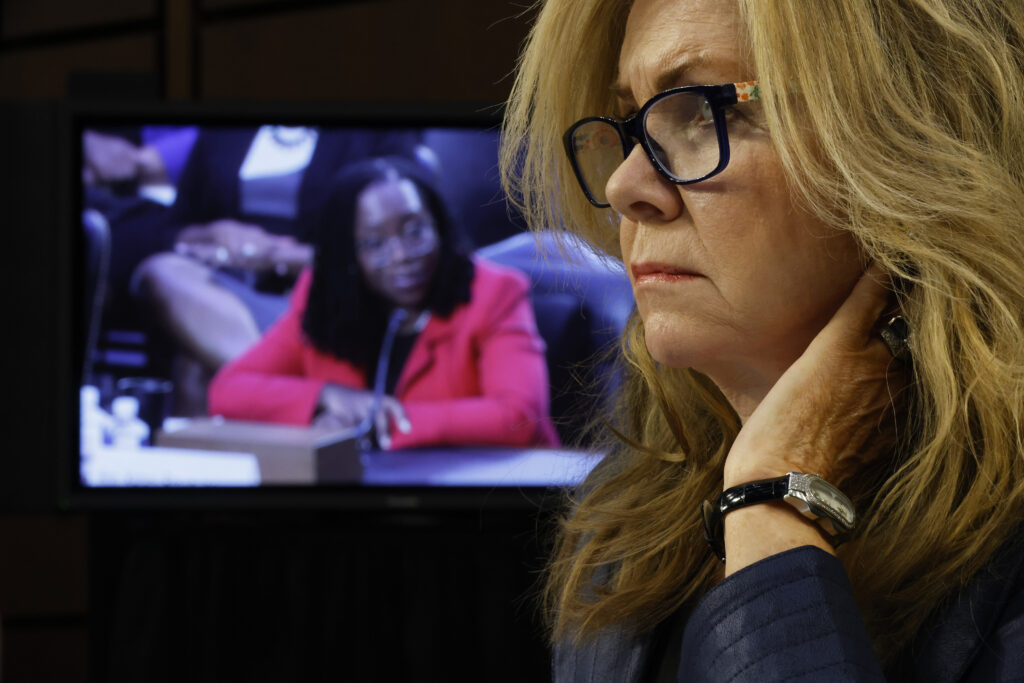 A blonde woman wearing glasses and a dark blue blazer sits in front of a television monitor showing a woman with dark, locked hair and a red blazer seated in a courtroom.