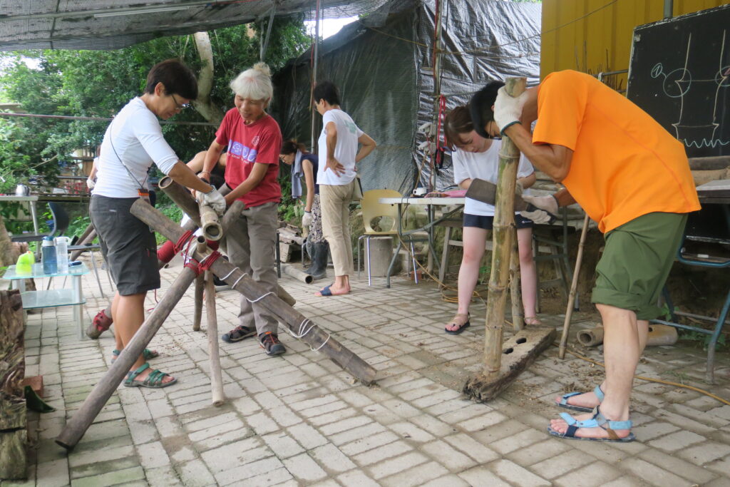Seven people work underneath a tarp to tie and assemble long pieces of bamboo with white rope.