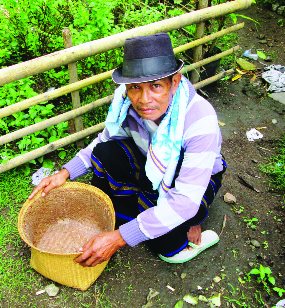 In front of a wooden fence, a person wearing a brimmed hat, scarf, and flip-flops crouches on the ground while holding a woven basket in front of him.