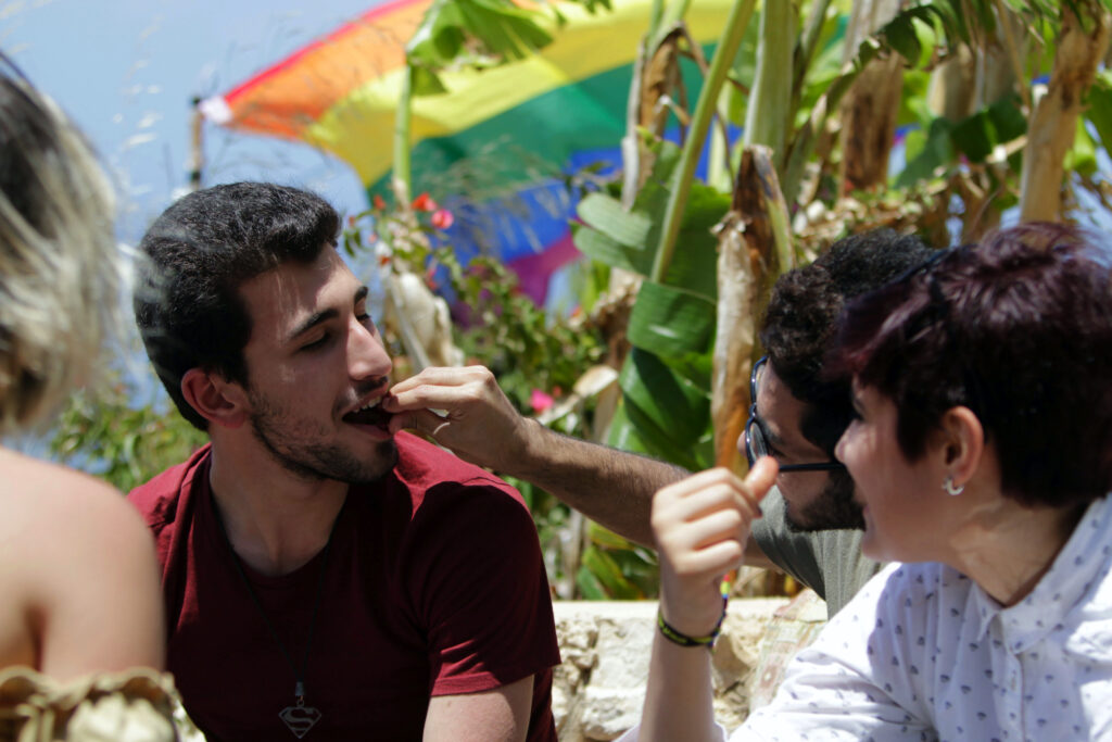 On a sunny day, four people sit outside. One bearded person wearing sunglasses feeds another bearded person as a rainbow flag is flying in the background.