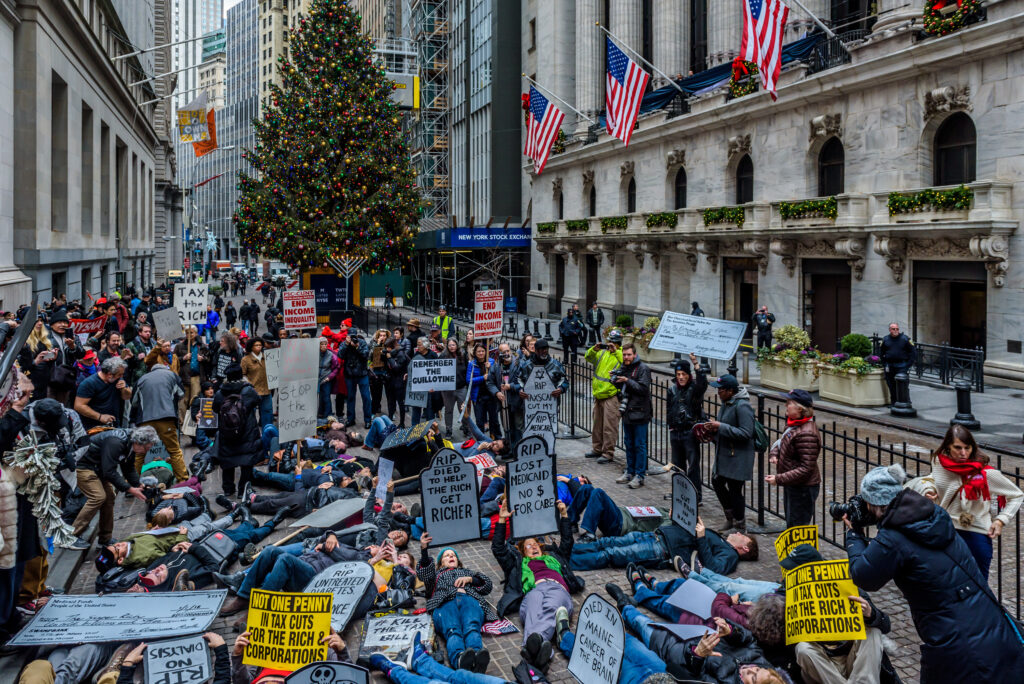 In a large city square, several people hold signs and raise fists while lying down on the ground. People stand around them, holding signs and taking pictures. Some signs read, “RIP, Died to help the rich get richer,” “RIP Lost Medicaid, no money for care,” “RIP, no dialysis,” “RIP Untreated Diabetes,” and “Tax the Rich.”