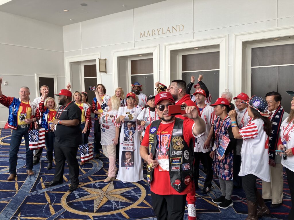In front of a wall inscribed with the word “Maryland,” people pose for pictures wearing red baseball caps with sayings such as “Trump Was Right” and “Trump Forever.” They wear clothing adorned with U.S. flags, an older man’s face, and similar phrases.